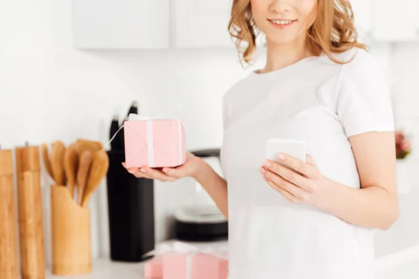 Cropped view of girl in pajamas with gift box using smartphone in kitchen — Stock Photo