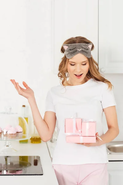 Bella ragazza sorpresa in pigiama e maschera addormentata contenente scatole regalo in cucina — Foto stock