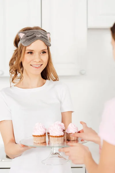 Selective focus of beautiful smiling girl in sleeping mask and pajamas holding glass stand with cupcakes — Stock Photo
