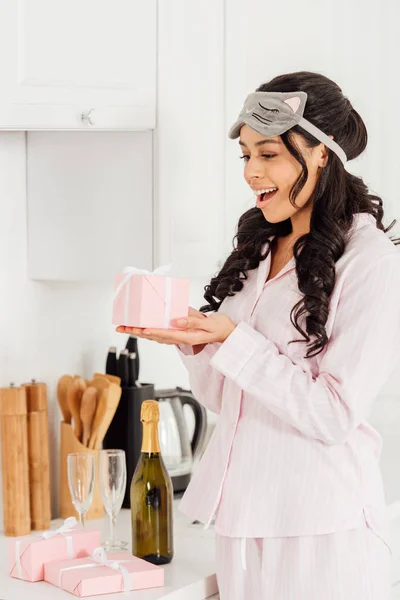 Hermosa chica afroamericana excitada en la máscara de dormir sosteniendo caja de regalo rosa en la cocina - foto de stock