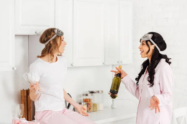 Beautiful multicultural girls talking and celebrating with champagne during pajama party in kitchen — Stock Photo