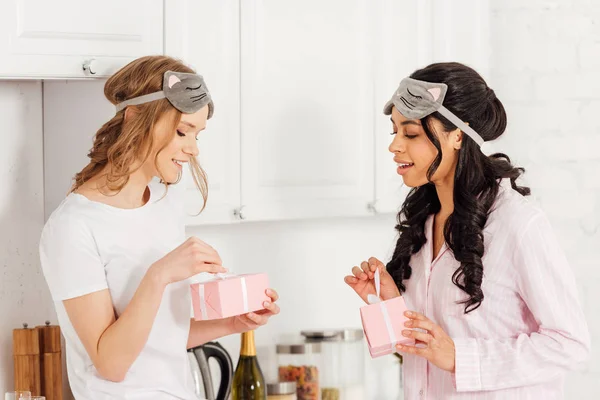 Beautiful multicultural girls in sleeping masks and pajamas opening gift boxes in kitchen — Stock Photo