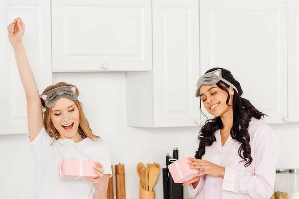 Belles filles multiculturelles heureuses en masques de sommeil et pyjamas avec des boîtes-cadeaux dans la cuisine — Photo de stock