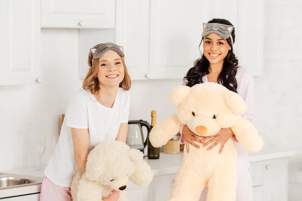 Hermosas chicas multiculturales con osos de peluche mirando a la cámara durante la fiesta de pijama en casa - foto de stock