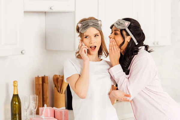 African american girl whispering secret to beautiful girl in sleeping mask talking on smartphone during pajama party — Stock Photo