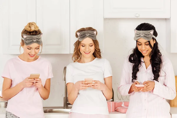Красивые улыбающиеся мультикультурные девушки в масках, используя смартфоны во время пижамной вечеринки — стоковое фото