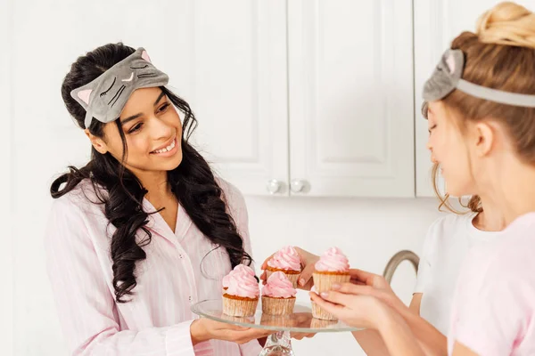 Beautiful multicultural girls in sleeping masks taking cupcakes from glass stand during pajama party — Stock Photo