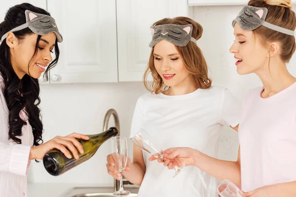 Красивые улыбающиеся мультикультурные девушки празднуют и разливают шампанское во время пижамной вечеринки — стоковое фото