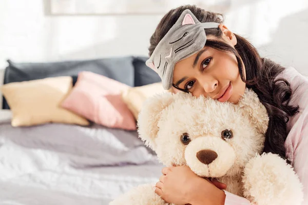 Beautiful african american girl in sleeping mask hugging teddy bear and looking at camera in bedroom — Stock Photo