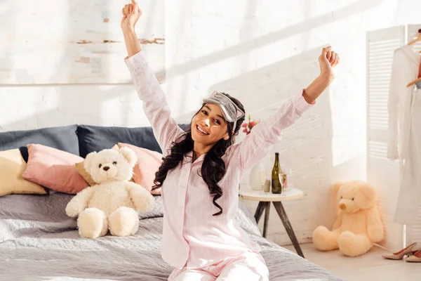 Hermosa chica afroamericana en la máscara de dormir sonriendo y estirándose en el dormitorio - foto de stock