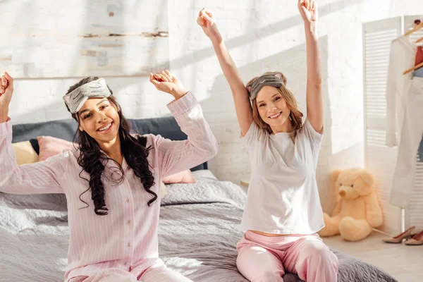 Hermosas chicas sonrientes multiculturales mirando a la cámara y estirándose en la mañana durante la fiesta de pijama - foto de stock