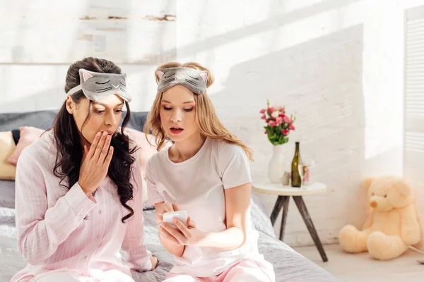 Красиві мультикультурні дівчата в сплячих масках сидять на ліжку і використовують смартфон під час вечірки піжами — стокове фото