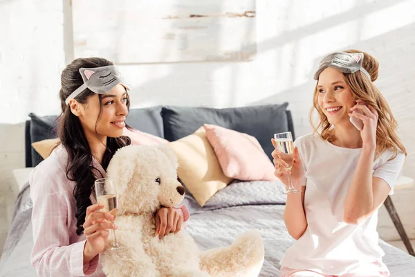 Schöne Mädchen in Schlafmaske spricht auf Smartphone, während afrikanisch-amerikanische Mädchen mit Champagnerglas während Pyjama-Party — Stockfoto