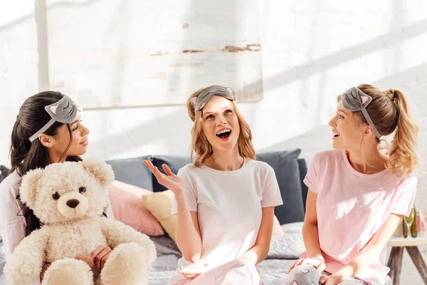 Hermosas chicas multiculturales en máscaras para dormir sentadas en la cama con oso de peluche y hablando durante la fiesta de pijama - foto de stock