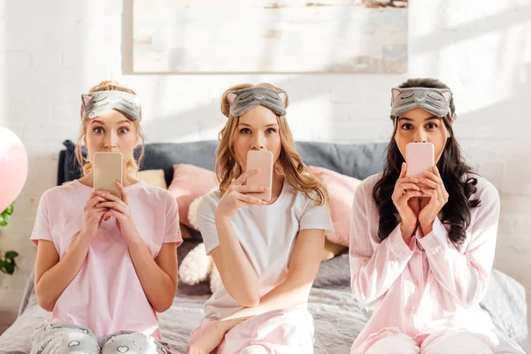 Beautiful multicultural girls in sleeping masks sitting on bed and covering mouths with smartphones during pajama party — Stock Photo