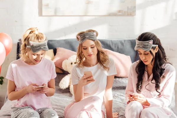 Красивые мультикультурные девушки в спальных масках сидят на кровати и используют смартфоны во время пижамной вечеринки — Stock Photo