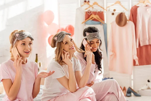 Красивые мультикультурные девушки в спальных масках сидят на кровати и разговаривают на смартфонах во время пижамной вечеринки — стоковое фото