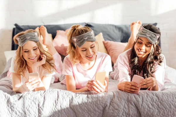 Hermosas chicas sonrientes multiculturales en máscaras para dormir tumbadas en la cama y usando teléfonos inteligentes durante la fiesta de pijama - foto de stock