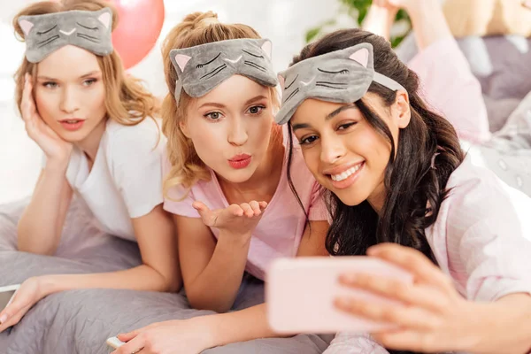 Красиві усміхнені мультикультурні дівчата в спальних масках лежать в ліжку і приймають селфі на смартфон під час вечірки піжами — стокове фото