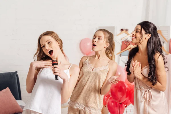 Hermosas chicas multiétnicas en ropa de dormir cantando karaoke en la fiesta de pijama en el dormitorio - foto de stock