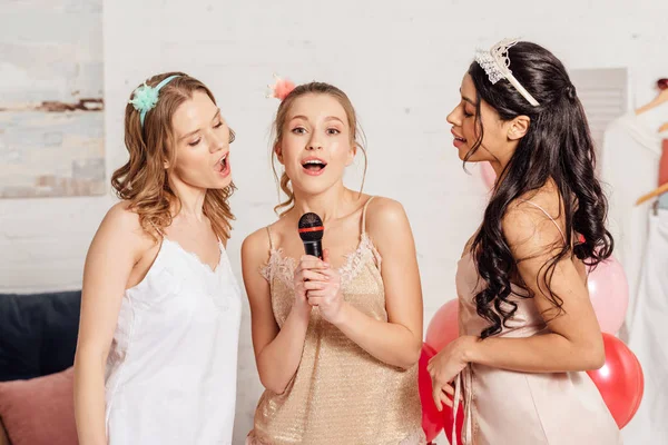 Schöne multiethnische Mädchen in Nachtwäsche und Stirnbändern singen Karaoke bei Pyjama-Party im Schlafzimmer — Stockfoto