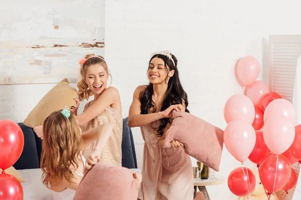Hermosas chicas multiétnicas en diademas divirtiéndose y luchando con almohadas durante la fiesta de pijama en el dormitorio - foto de stock