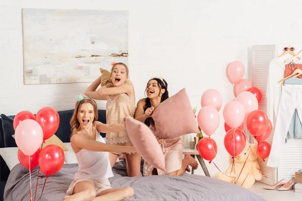 Beautiful multiethnic girls in nightwear having fun and fighting with pillows during pajama party in bedroom — Stock Photo