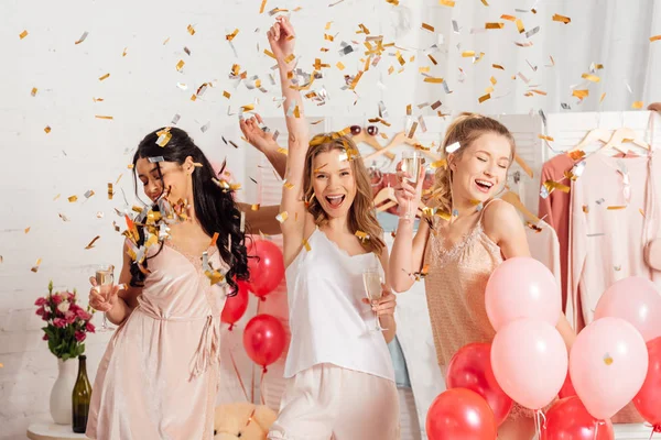 Beautiful cheerful multicultural girls holding champagne glasses and celebrating under falling confetti during pajama party — Stock Photo
