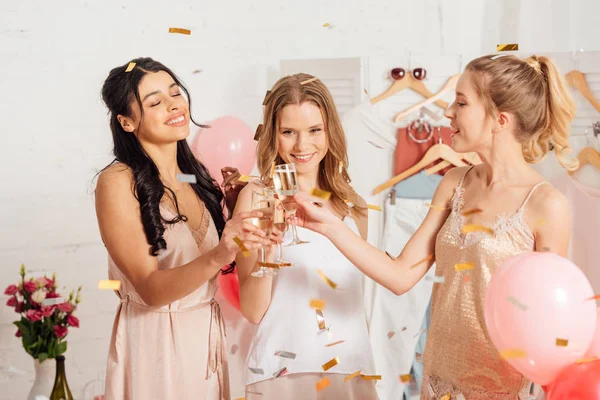 Meninas multiculturais bonitas clinking taças de champanhe e celebrando sob queda confetti durante a festa de pijama — Fotografia de Stock