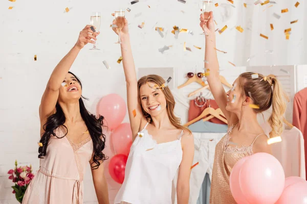 Beautiful multicultural girls toasting with champagne glasses and celebrating under falling confetti during pajama party — Stock Photo