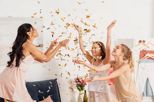 Hermosas chicas multiculturales en ropa de dormir que se divierten bajo la caída de confeti durante la fiesta de pijama - foto de stock