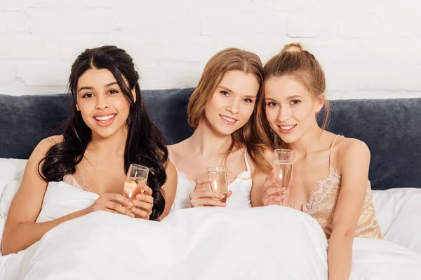 Schöne multikulturelle Mädchen mit Champagnergläsern im Bett, die während einer Pyjama-Party in die Kamera schauen — Stockfoto
