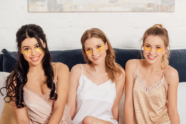 Belle ragazze multiculturali sorridenti con patch occhi dorati guardando la fotocamera a letto durante il pigiama party — Foto stock