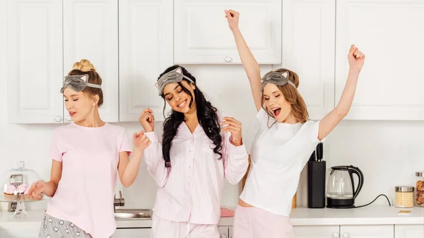 Hermosas chicas multiculturales en máscaras para dormir bailando y divirtiéndose en la cocina durante la fiesta de pijama - foto de stock
