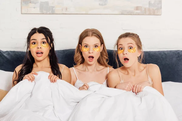 Красивые удивленные мультикультурные девушки с золотыми повязками на глазах, смотрящие на камеру в постели во время пижамной вечеринки — стоковое фото