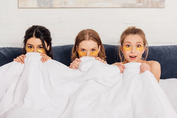 Hermosas chicas multiculturales con parches de ojos dorados mirando a la cámara en la cama durante la fiesta de pijama - foto de stock