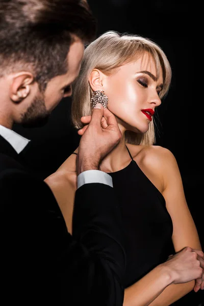 Hombre tocando elegantes pendientes de mujer atractiva aislado en negro - foto de stock