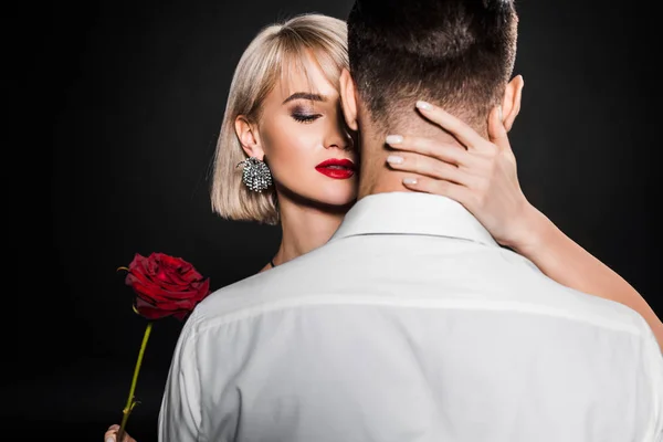 Leidenschaftliche Frau hält Rosenblume und umarmt Mann, isoliert auf schwarz — Stockfoto