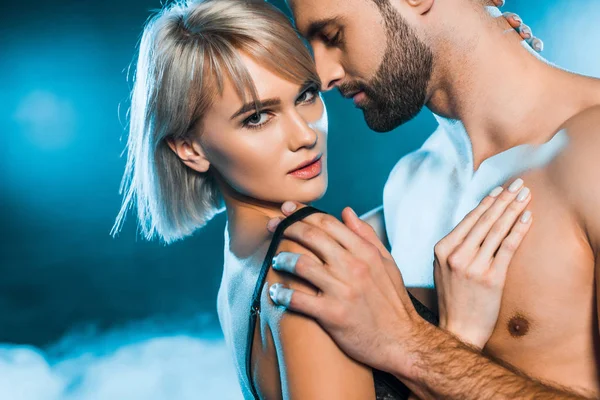 Uomo barbuto spogliarsi seducente giovane donna su sfondo blu fumoso — Foto stock