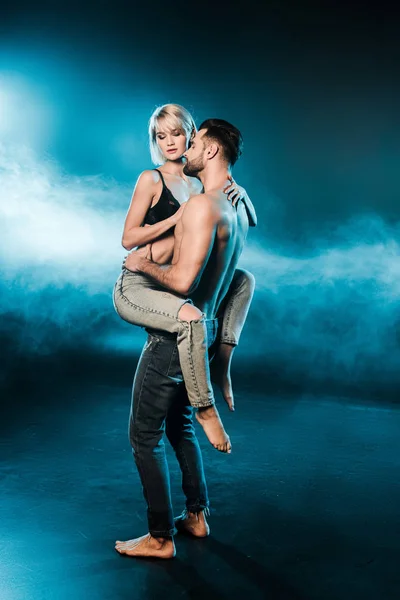 Красивий спокусливий чоловік тримає біляву дівчину на синьому димчастому фоні — стокове фото