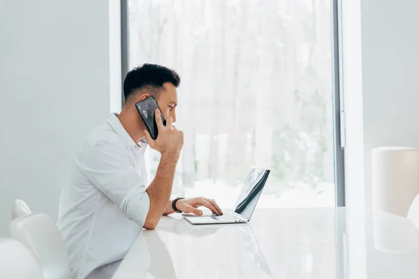 Homem ocupado falando no smartphone e digitando no teclado do laptop — Fotografia de Stock