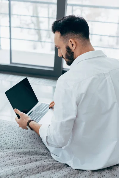 Брюнетка людина з бородою, сидячи на ліжку і використання ноутбука з порожній екран — стокове фото