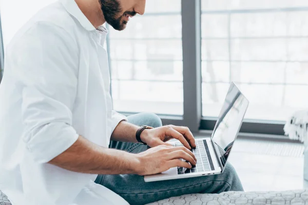 Vista parcial del hombre en camisa blanca y jeans escribiendo en el teclado del ordenador portátil - foto de stock