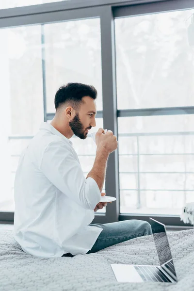 Чоловік у білій сорочці сидить на ліжку і п'є каву — стокове фото