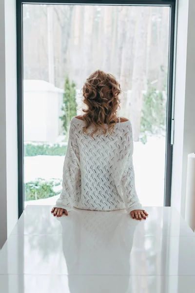 Vue arrière de la femme bouclée en pull tricoté regardant la fenêtre — Photo de stock