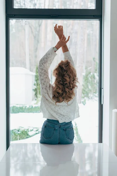 Visão traseira da mulher encaracolada de pé com as mãos para cima na frente da janela — Fotografia de Stock