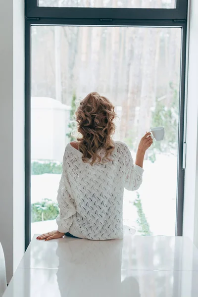 Vista posteriore della ragazza riccia in piedi davanti alla finestra con tazza di caffè — Foto stock