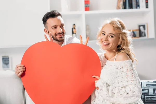 Cheerful couple holding red heart and showing peace sign — Stock Photo