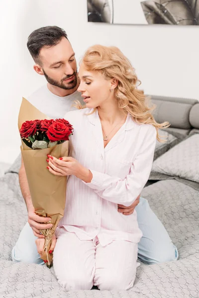 Uomo che presenta fiori alla fidanzata mentre seduto sul letto — Foto stock