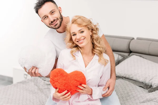 Excited couple holding toy hearts while sitting on bed — Stock Photo